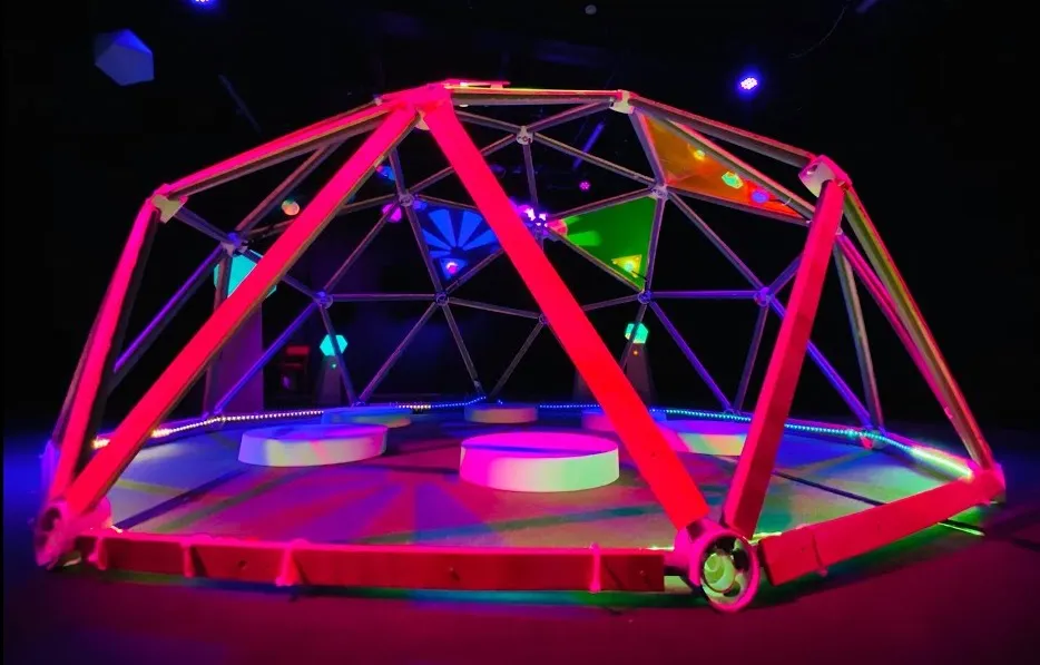 The finished dome at night, its frame lit pink and red, round play stations on the floor inside
