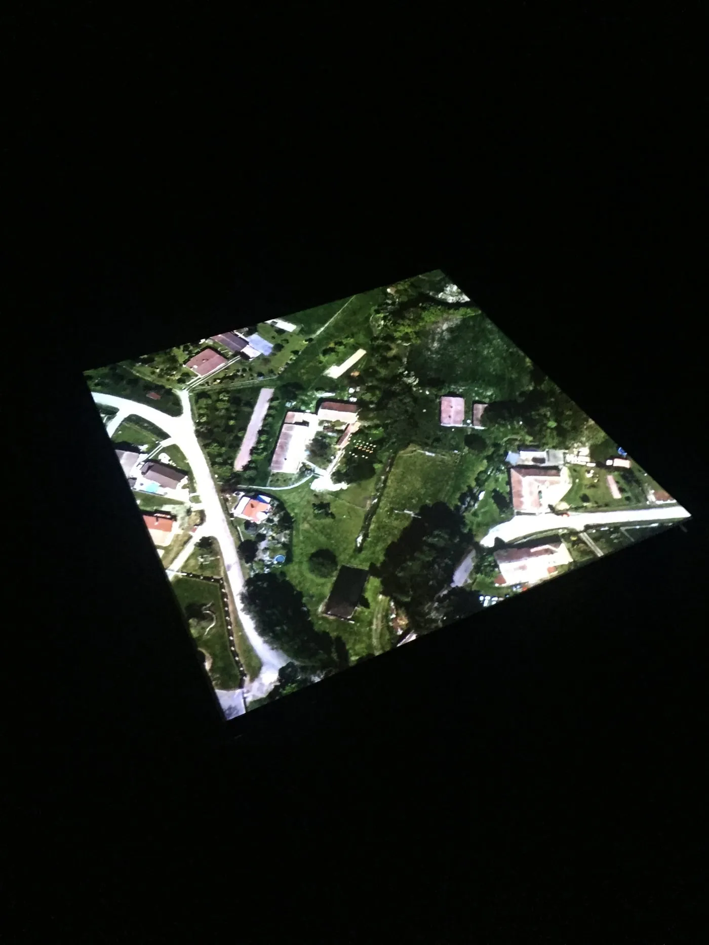A single square of satellite imagery projected onto the floor of a dark room: houses, gardens and a winding road seen from above, the rest of the room black