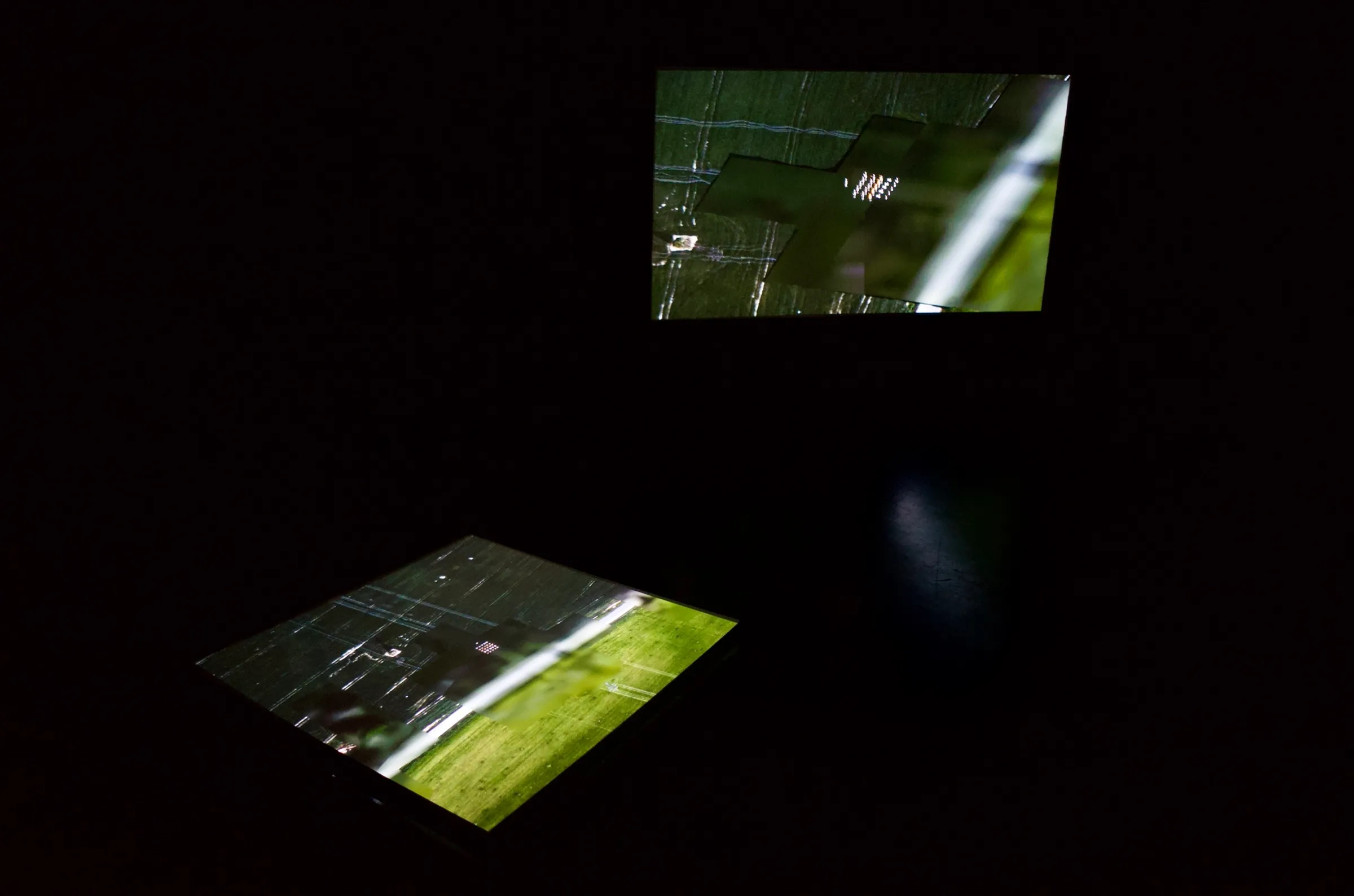 The installation in a dark room: one projection lying on the floor seen from above, a second hanging on the wall, both showing the same satellite terrain
