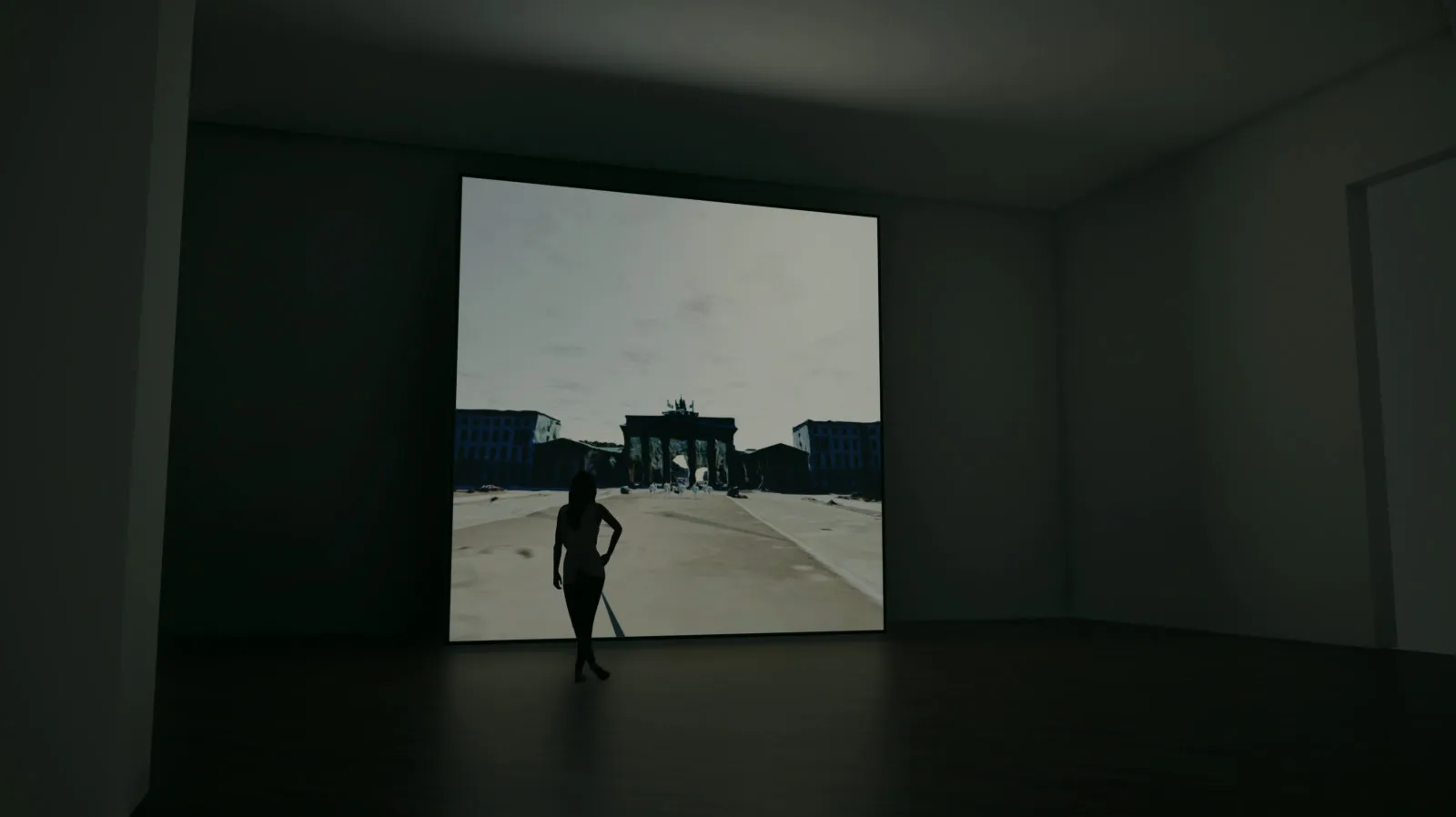 A dark room, one wall filled by a square screen showing a street-level view of the Brandenburger Tor, a woman standing before it in silhouette
